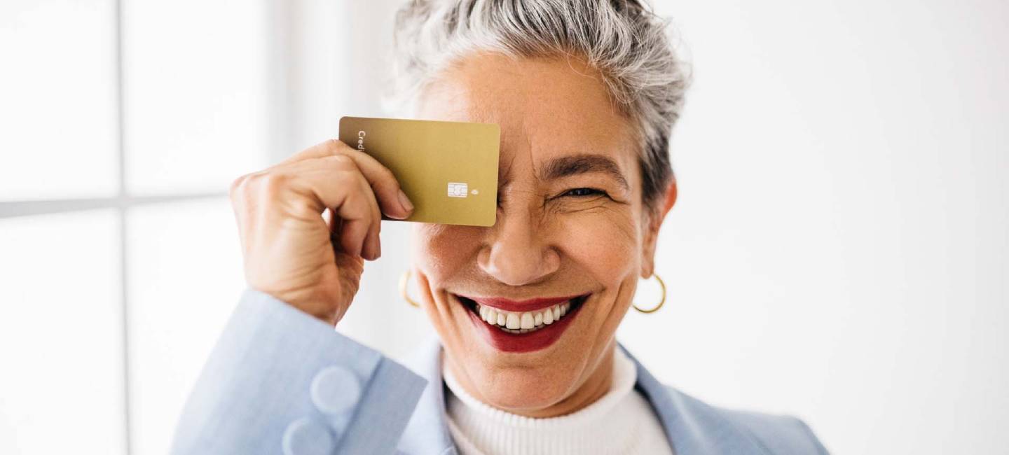 A mature woman in a blue blazer holding up a gold credit card over her eye