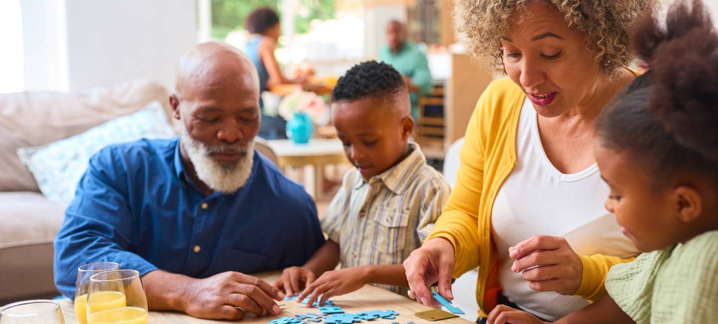 Grandparents With Grandchildren Indoors At Home Doing Jigsaw Puzzle With Parents In Background