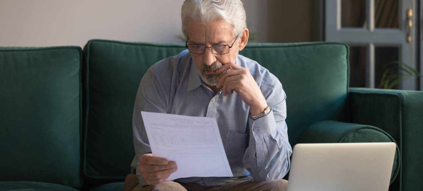 Mature man wearing glasses sitting on couch in living room manage budget received invoice analyses month expenses feels concerned about council tax letter