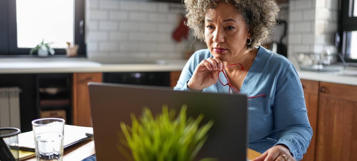 a concerned mature woman with curly gray hair reviews paperwork at her kitchen table she holds a document while looking at it intently, surrounded by a laptop