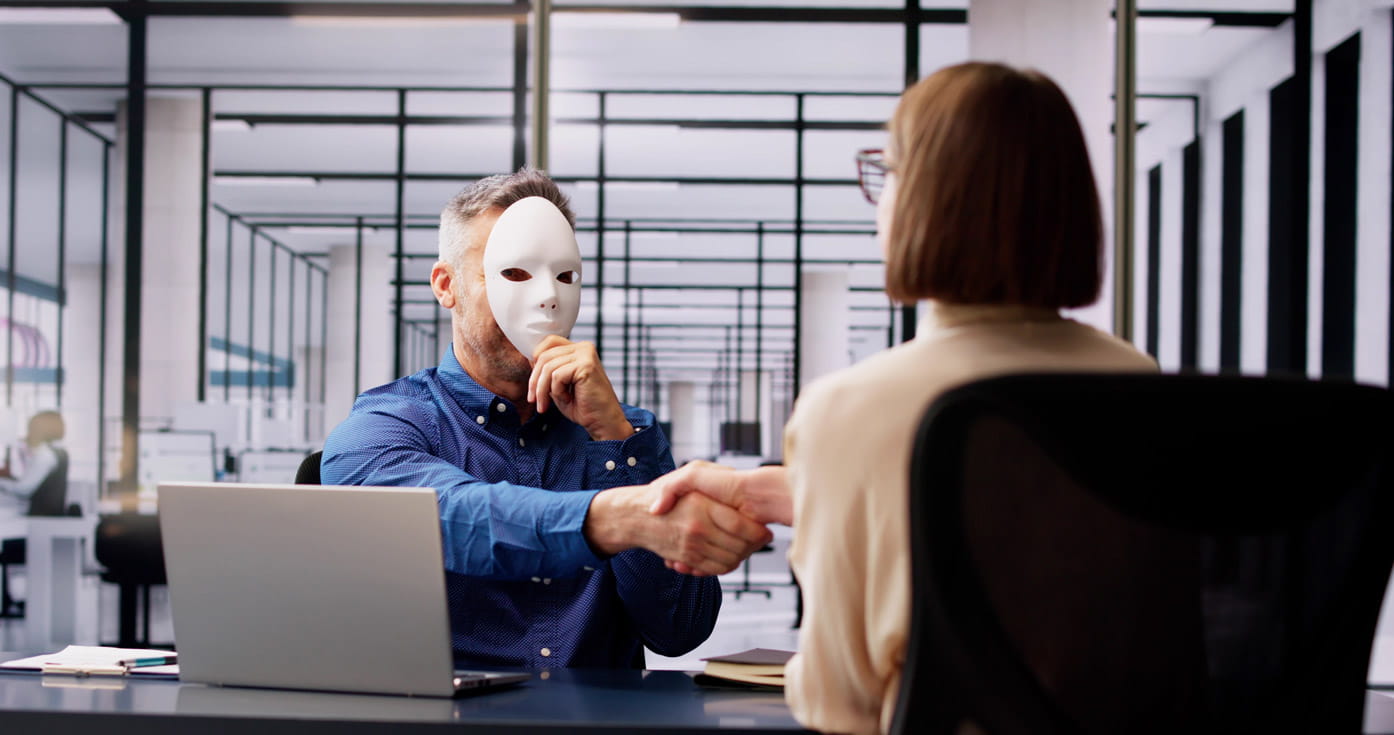 An untrustworthy man holding a white mask up to his face shakes a woman's hand over a desk
