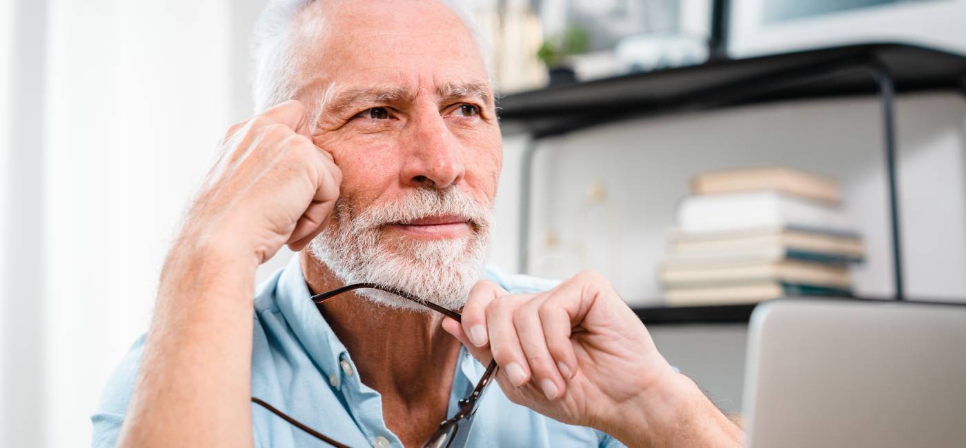 Tired mature 60s man working at laptop from home, taking off glasses, touching head and eyes. 