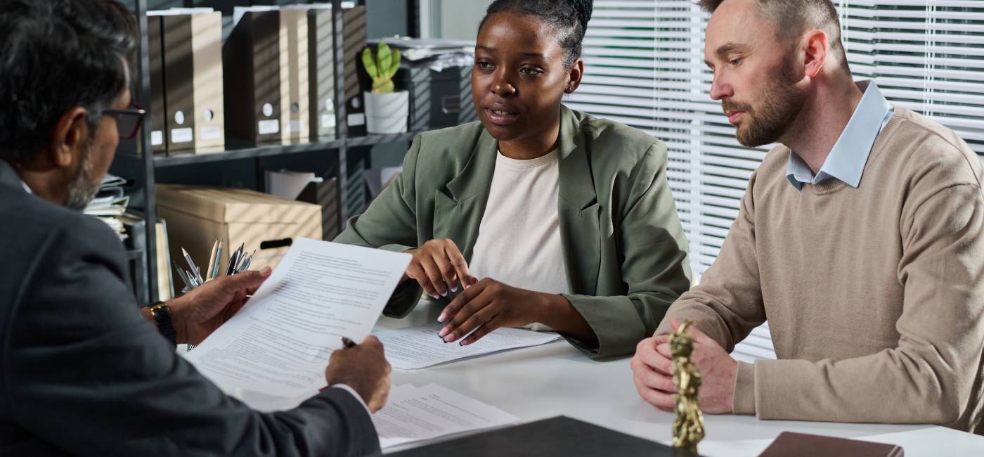 Young intercultural couple looking at mature male solicitor or notary with contract in hands sitting by desk in front of them
