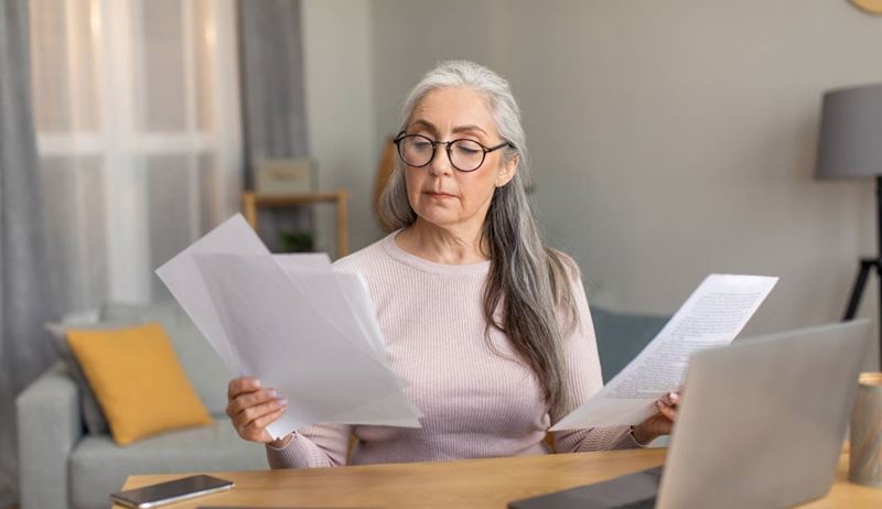 female with gray hair in glasses works on computer and documents in room interior. Bookkeeping, paying bills