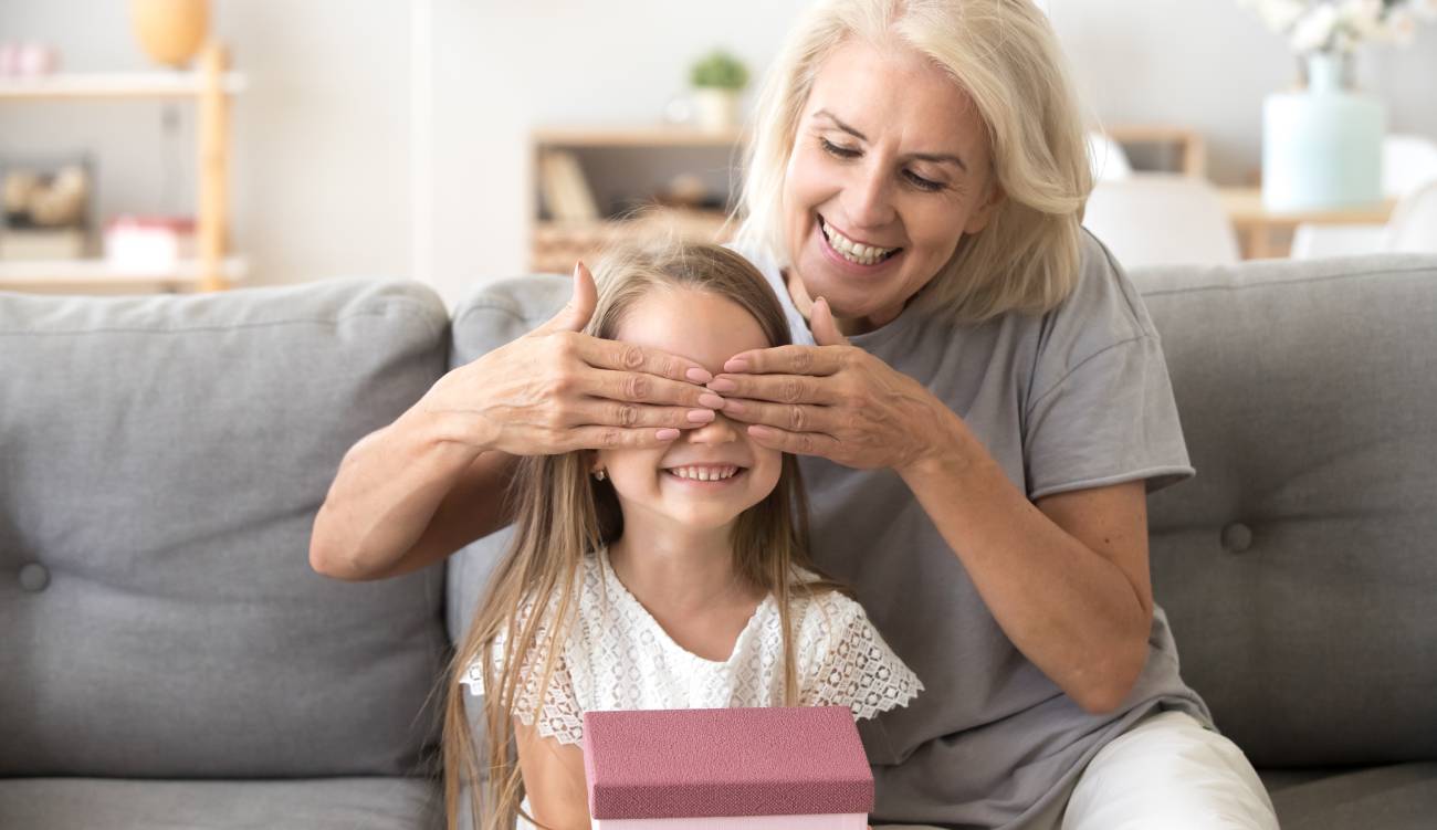 Loving old grandmother making present to little happy kid girl holding gift box, middle-aged granny closing eyes of cute smiling grandchild excited with birthday surprise from grandma sitting on sofa
