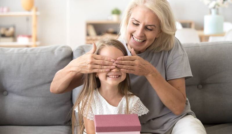 Loving old grandmother making present to little happy kid girl holding gift box, middle-aged granny closing eyes of cute smiling grandchild excited with birthday surprise from grandma sitting on sofa