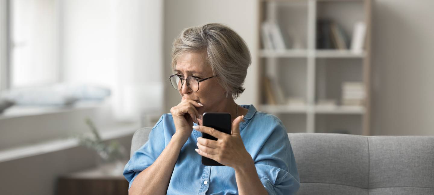 Unhappy older woman sitting on couch, holding smartphone feels frustrated and concerned, received upsetting text message.