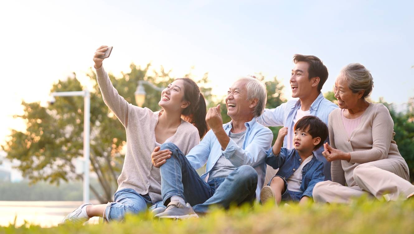 three generation happy asian family sitting on grass taking a selfie using mobile phone outdoors in park