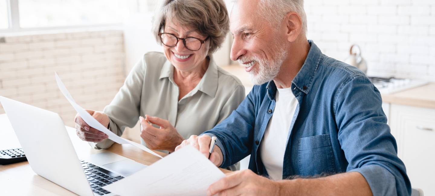 Smiling elderly senior couple husband and wife using laptop and doing paperwork together at home kitchen.