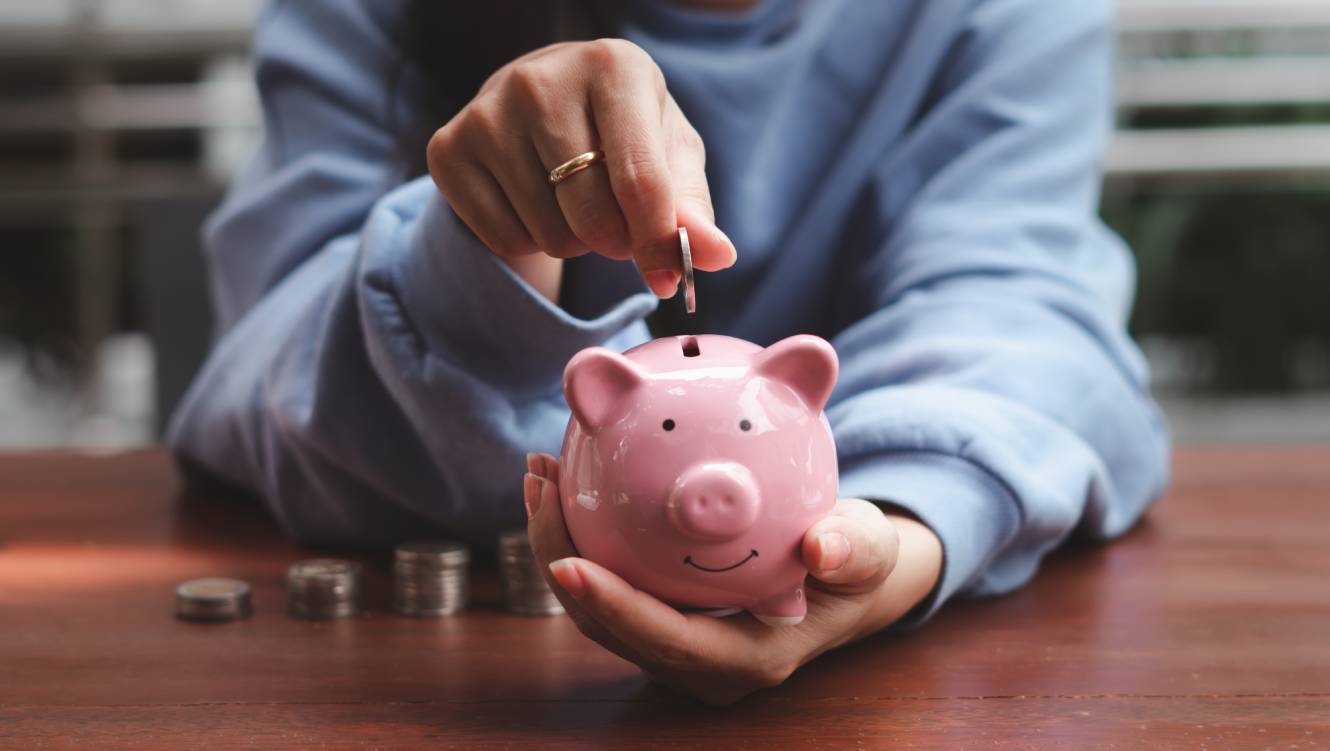 Womans hand with a gold ring on her middle finger putting a coin in to a piggy bank