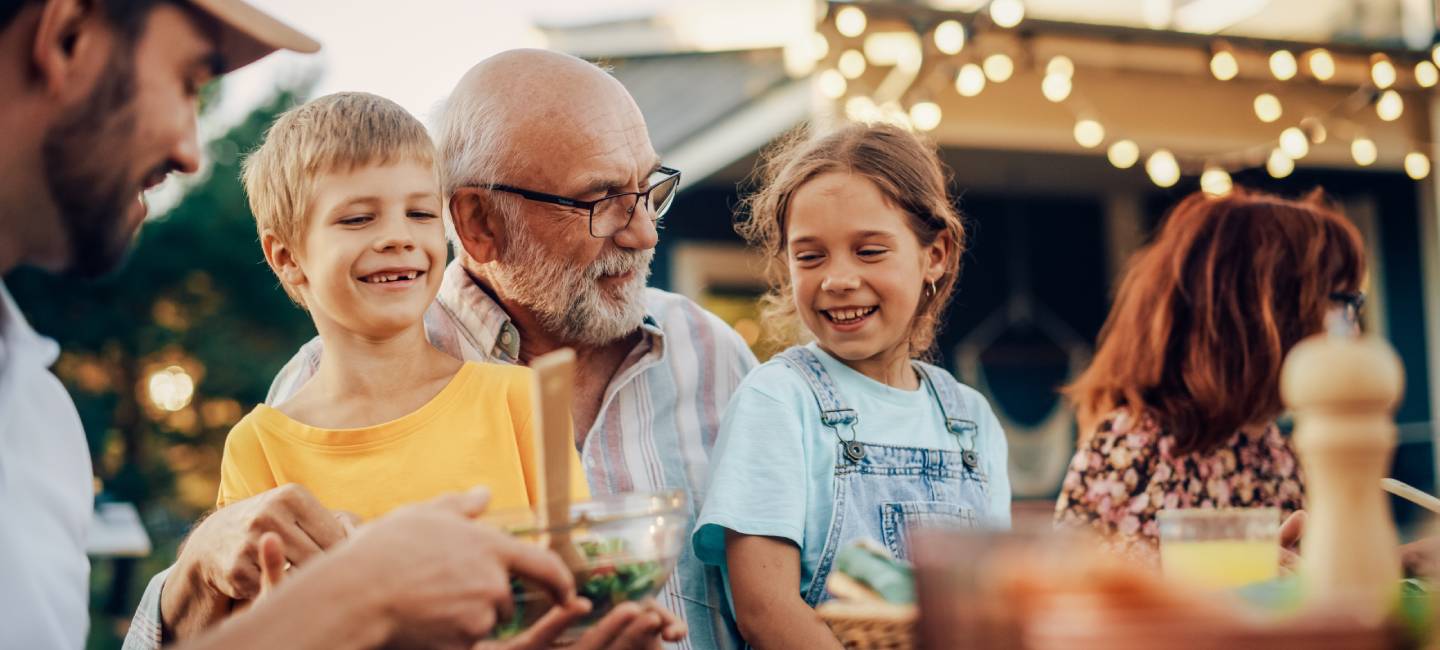 Happy Senior Grandfather Talking and Having Fun with His Grandchildren, Holding Them on Lap at a Outdoors Dinner with Food and Drinks. 