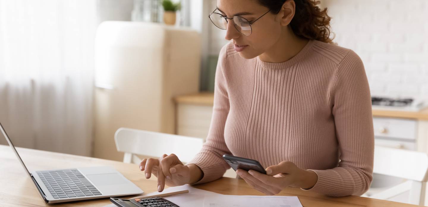 Concentrated millennial woman in eyeglasses managing monthly budget, calculating medical insurance, planning investments, paying utility household bills or taxes online using mobile phone application.