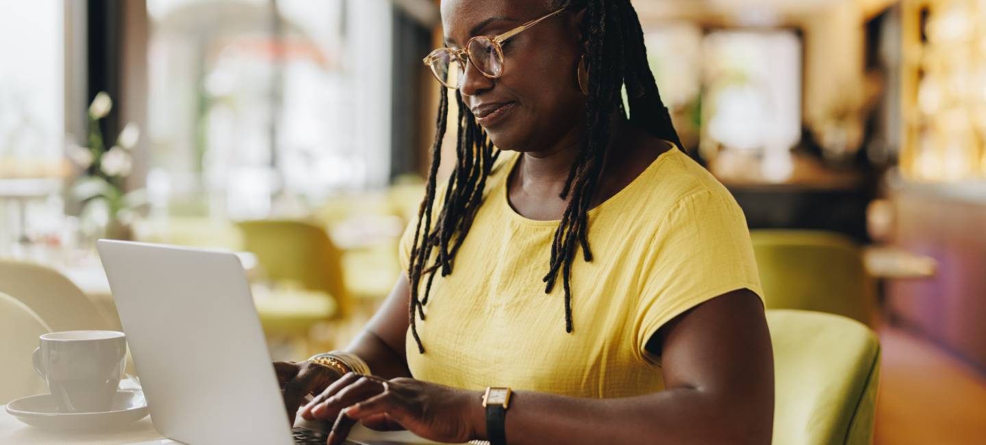 Self-employed senior woman typing an email on a laptop while working in a cafe. Mature entrepreneur with dreadlocks doing some online freelance work.
