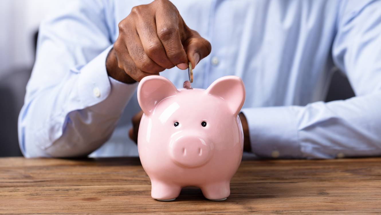 Man's Hand Inserting Coin In Piggybank Over Wooden Desk