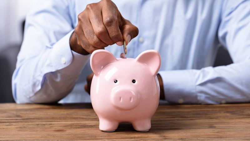 Man's Hand Inserting Coin In Piggybank Over Wooden Desk