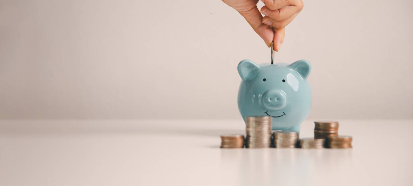 A hand putting copper coins in to a blue piggybank, with copper coins stacked in front of the piggybank