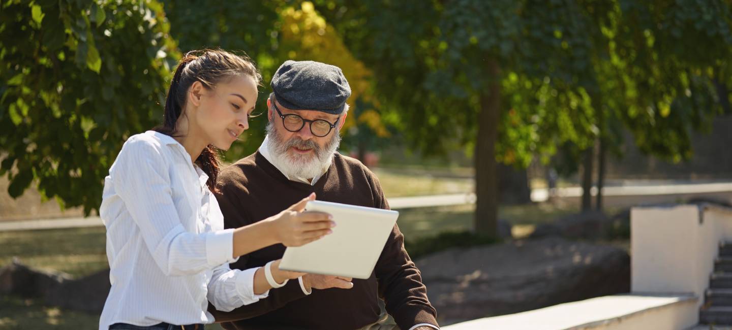 A young smiling girl sitting with grandfather and laptop in a park.