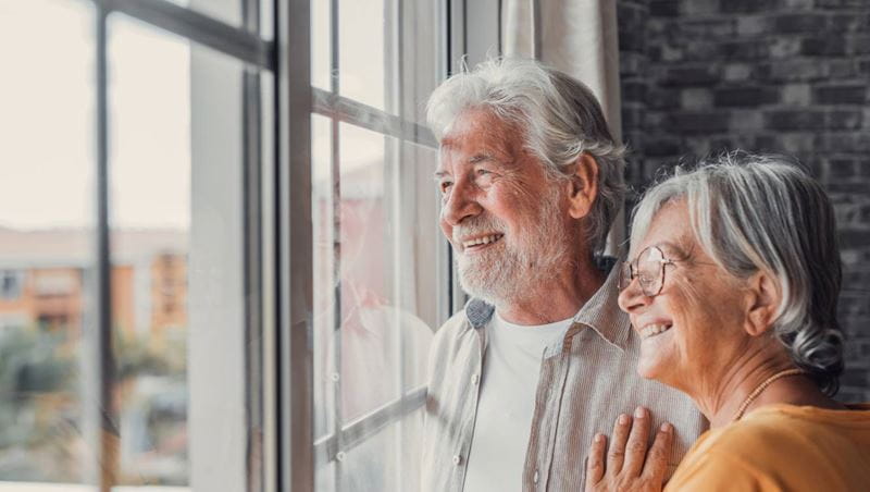 Happy bonding loving middle aged senior retired couple standing near window, looking in distance, recollecting good memories or planning common future, enjoying peaceful moment together at home.