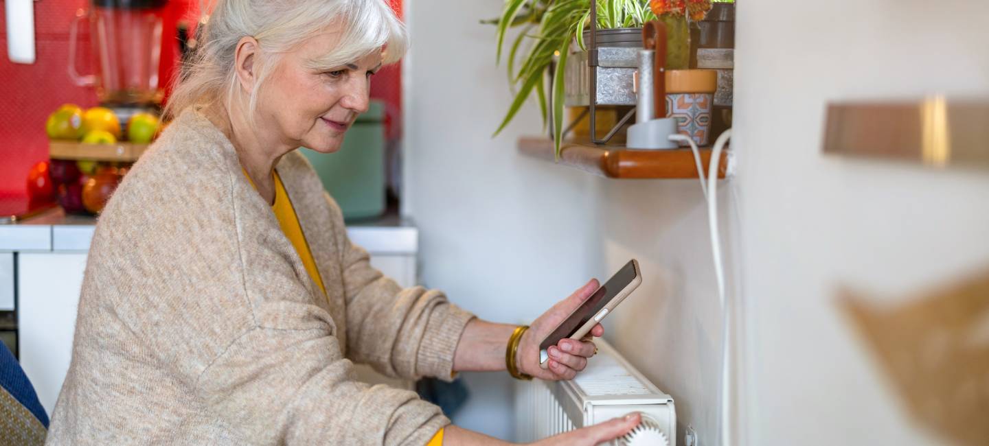 A mature woman holding mobile phone while adjusting the thermostat on the radiator at home