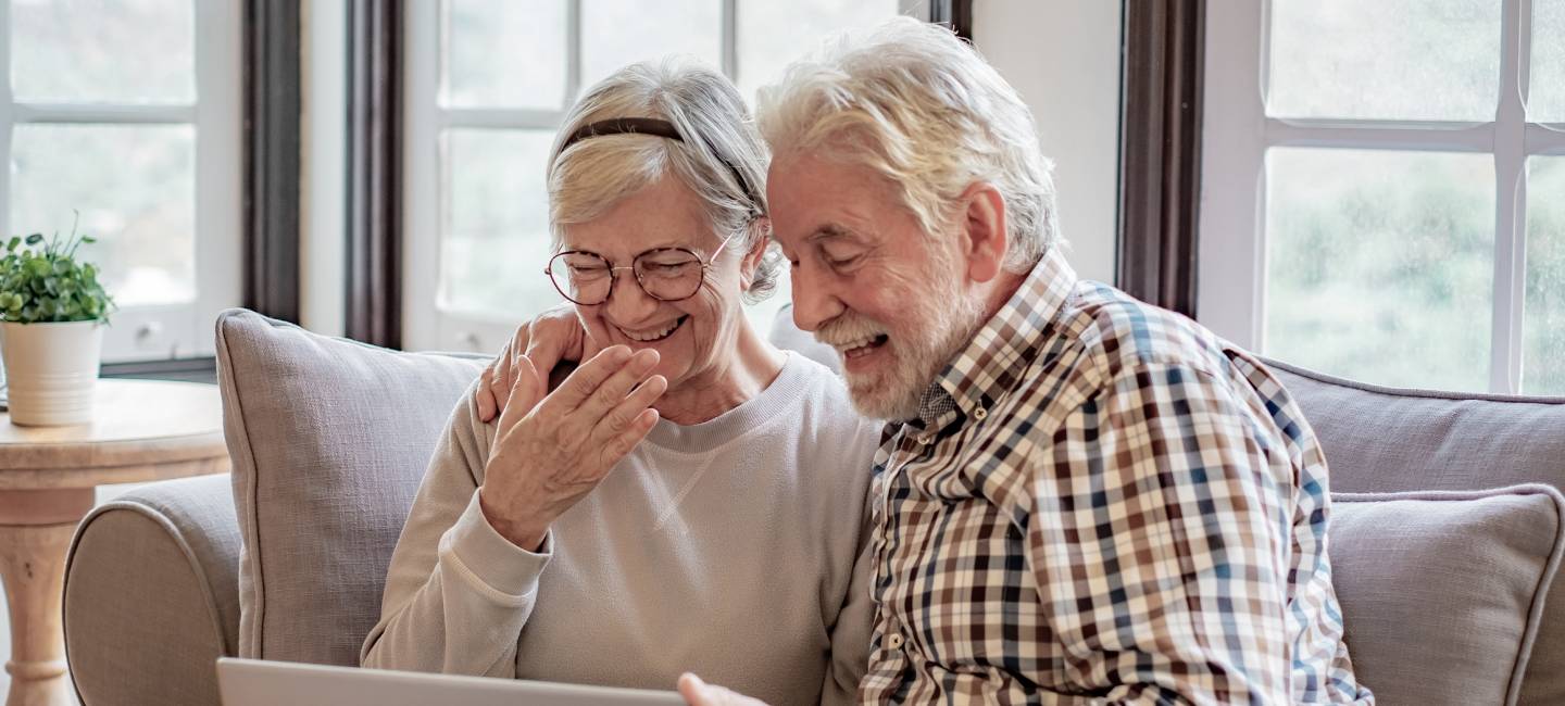 Portrait of cheerful senior couple embracing while sitting on sofa laughing. Elderly happy couple relaxing while browsing together on laptop