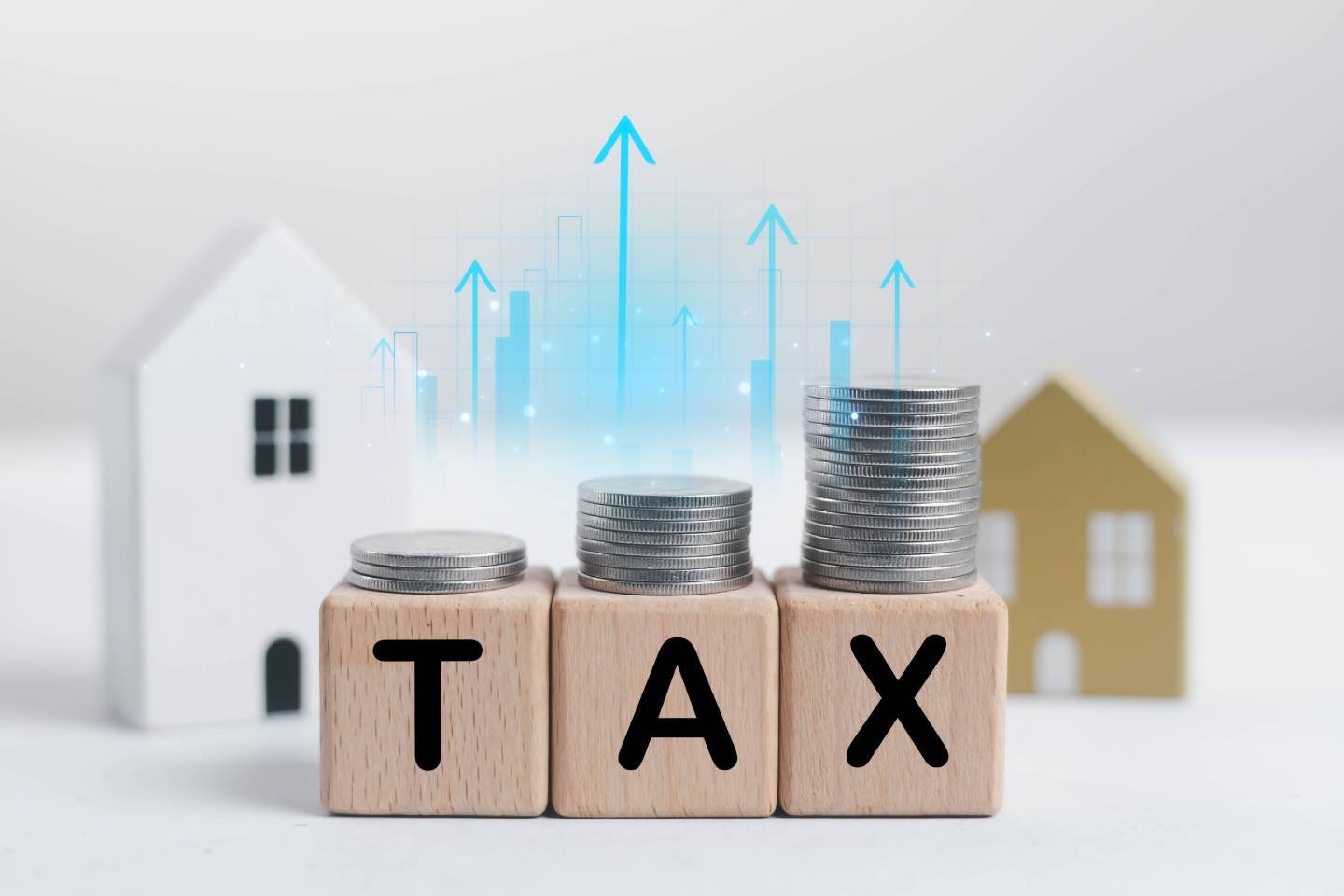  Building bocks with the letters TAX on them with coins sitting at the top of the building blocks with blue arrows pointing up with house modules in the back