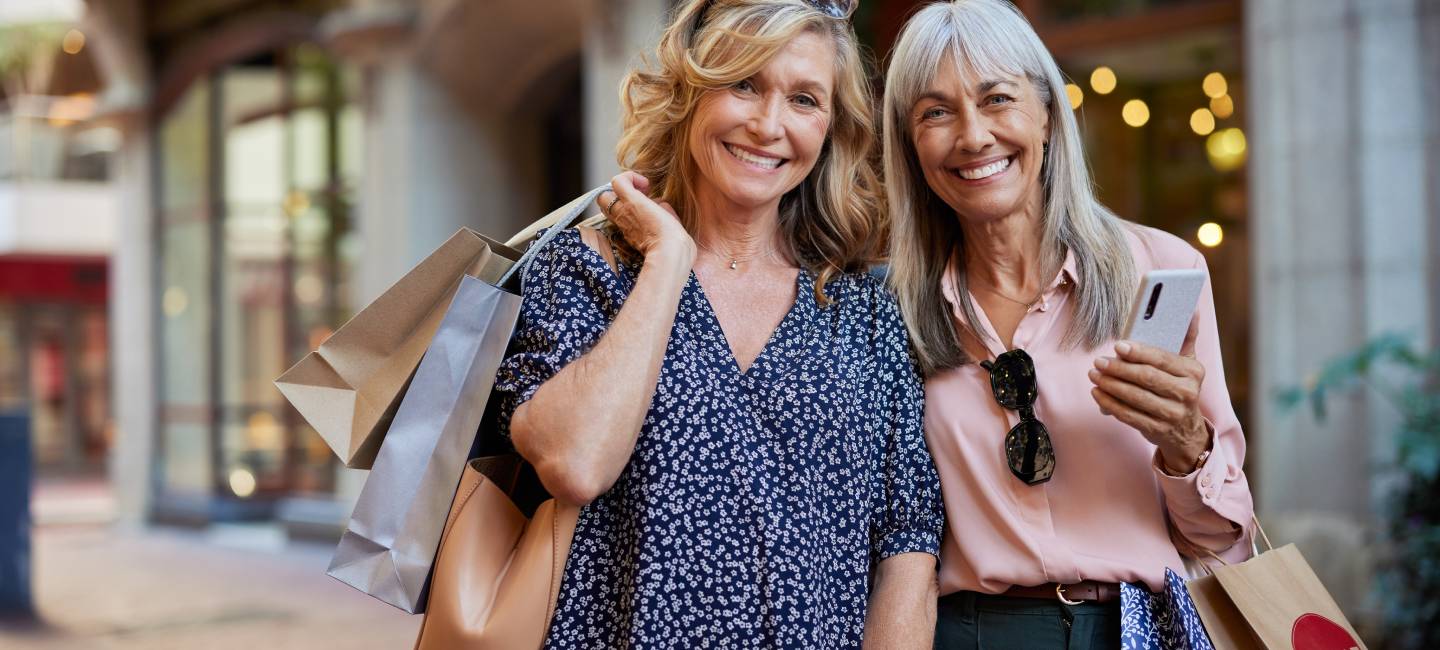 Two smiling mature ladies pose happily while holding shopping bags during sale season