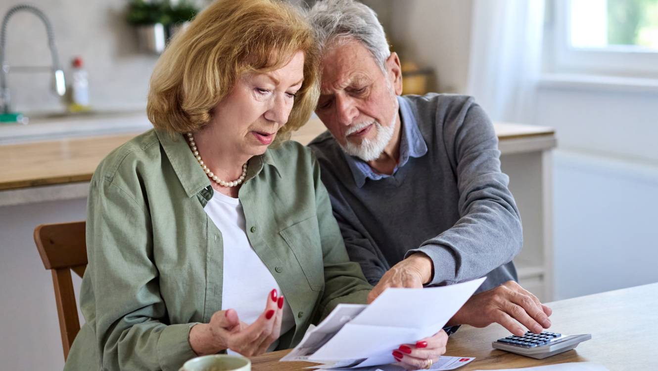 Senior couple checking bills and calculating expenses using a calculator at home, debt taxes and home budget concept, worried married elderly couple