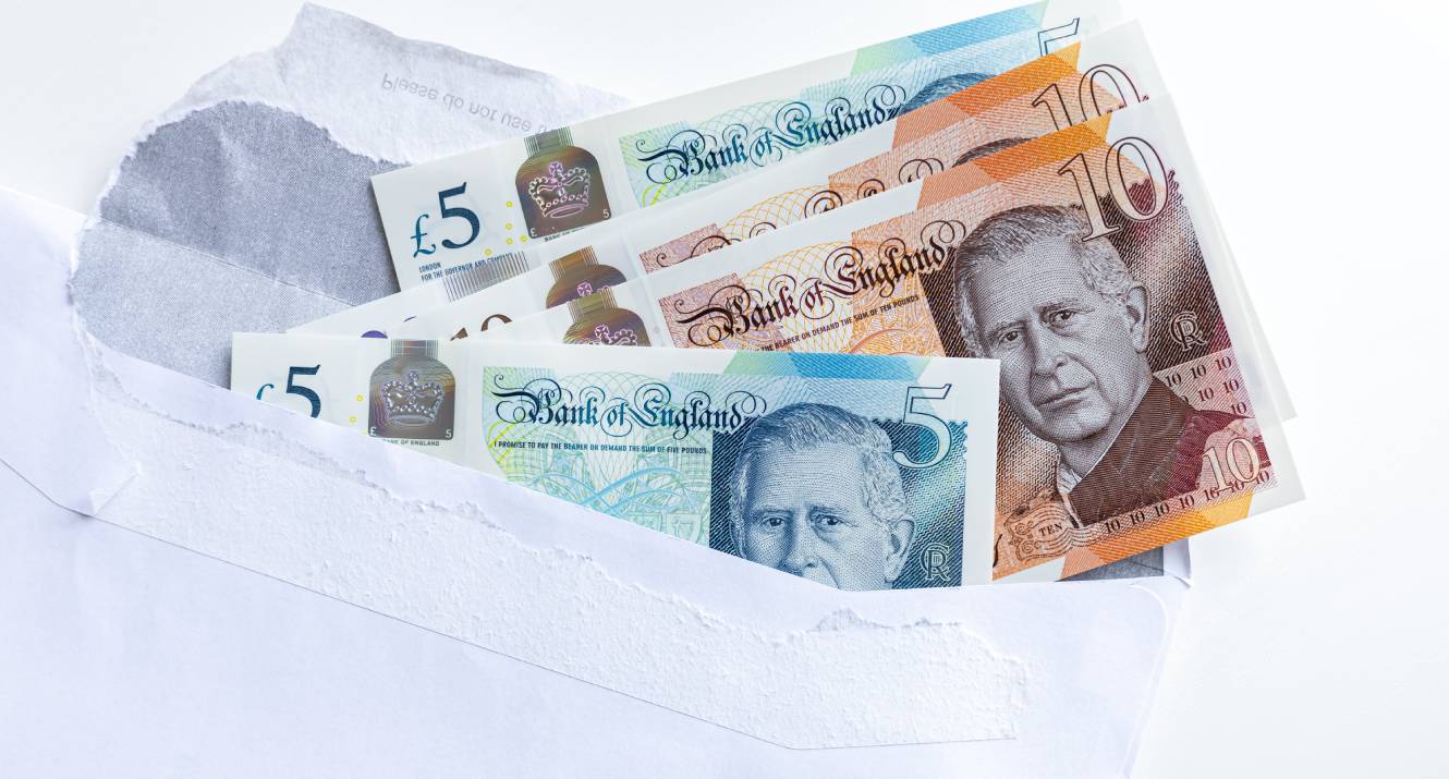 British pound sterling banknotes with King Charles portrait tucked in a white envelope on wooden desk