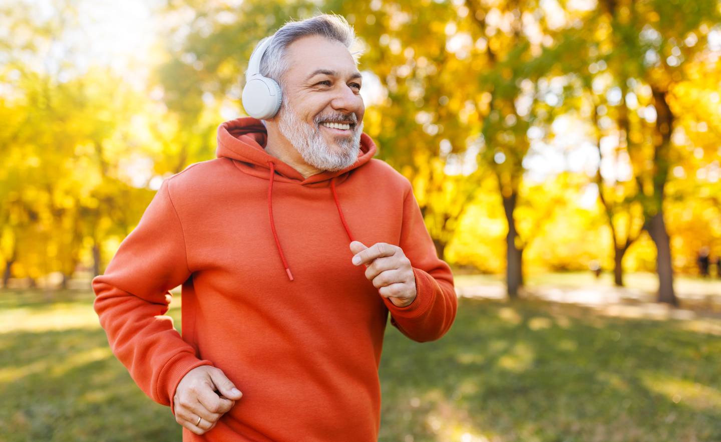 Happy senior man in headphones and sportswear jogging running outside in autumn park. 
