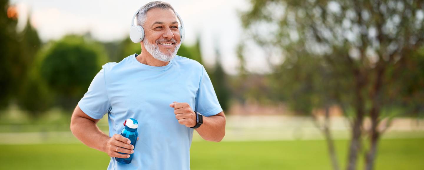 Happy mature man in headphones holding water bottle while jogging outside in park in early morning,