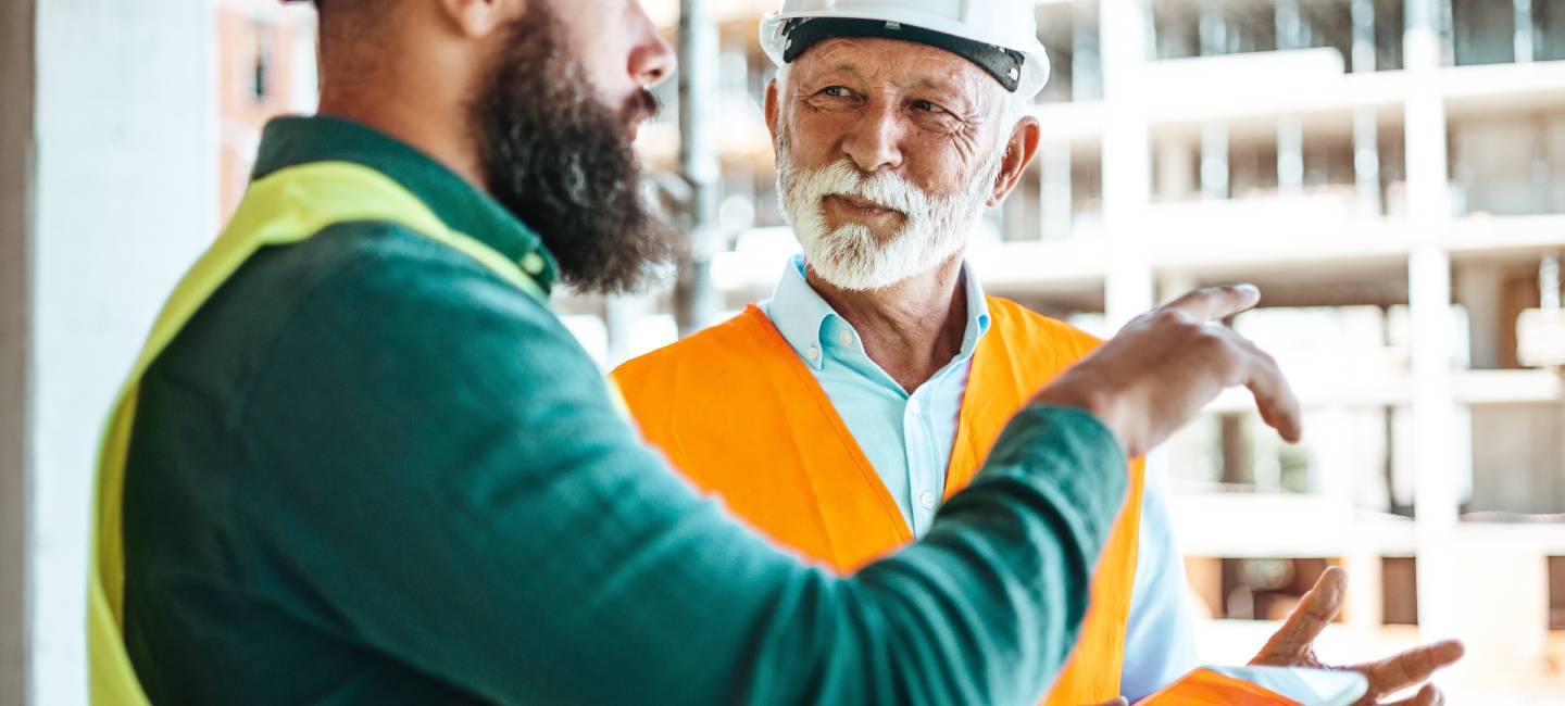 A younger worker in a green vest and hard hat shows a tablet to an older site manager in white, both engaged in active dialogue at a bustling construction location.