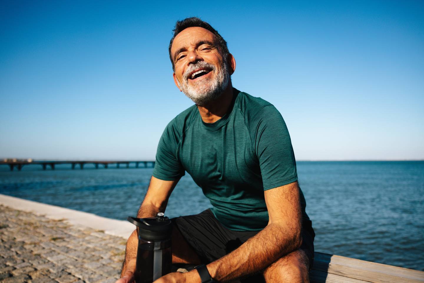 Mature male sitting by the water, smiling, enjoying nature during a sunny day outdoors.