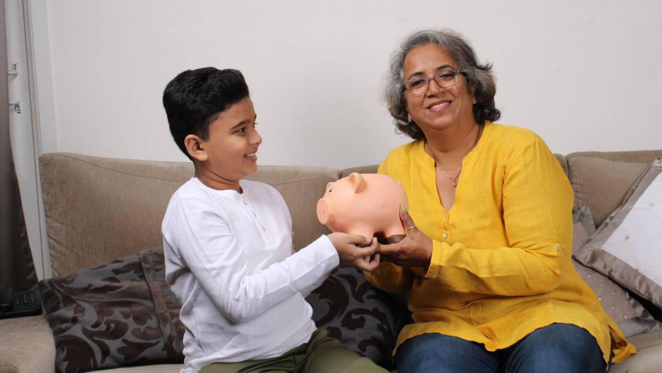 Grandma teaching her grand son importance of savings, putting coins into pink piggy bank.