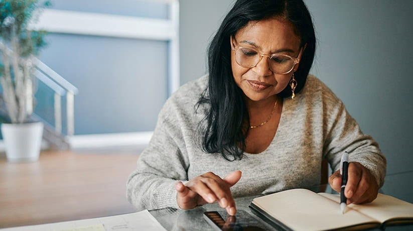 A woman writes in a diary while using a mobile phone to calculate expenses.