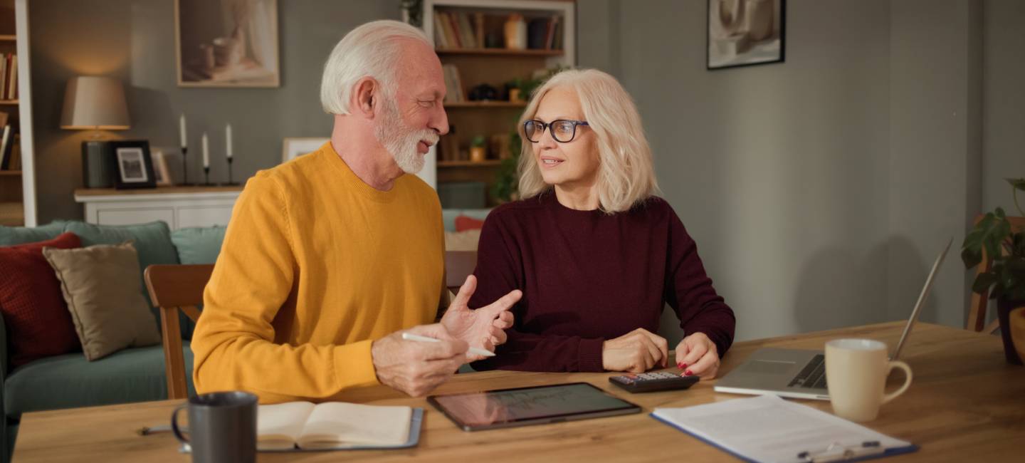 An older couple sits at a wooden table in their home discussing life insurance. They have financial documents, a tablet, and a calculator visible while they talk together.