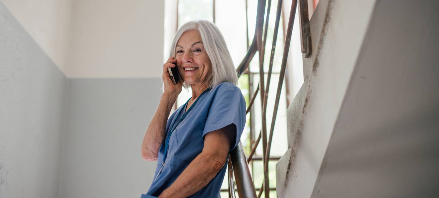 Mature NHS Nurse standing in a stairwell, on the phone, smiling. 