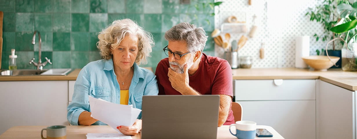 Mature husband and wife paying bills and managing family finances together on a laptop, discussing expenses in a cozy home environment.