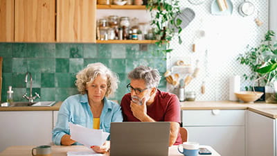 Mature husband and wife paying bills and managing family finances together on a laptop, discussing expenses in a cozy home environment.
