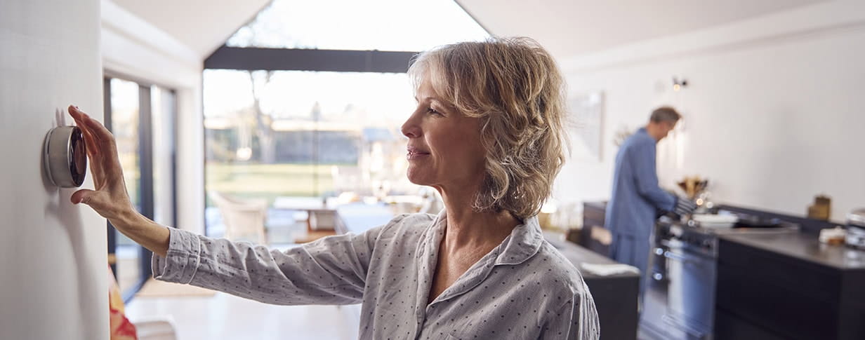 Mature woman adjusting her central heating thermostat control in her kitchen at home