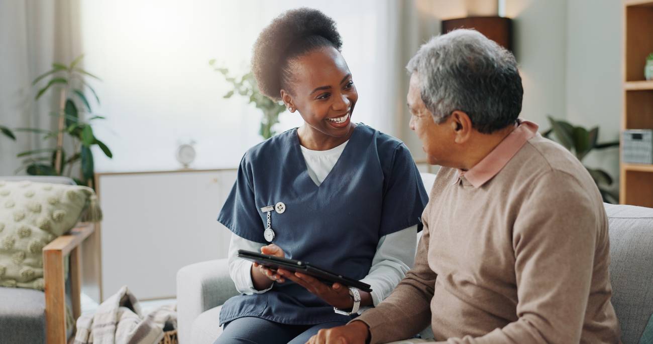 A young nurse smiling at an elderly man as they sit on the sofa looking at a tablet