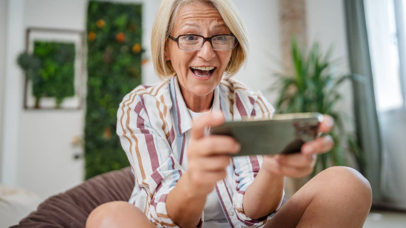 One mature blonde caucasian woman sit indoor happy smile play video games leisure activity at home or work hold mobile phone use smartphone have fun online copy space wear eyeglasses and shirt