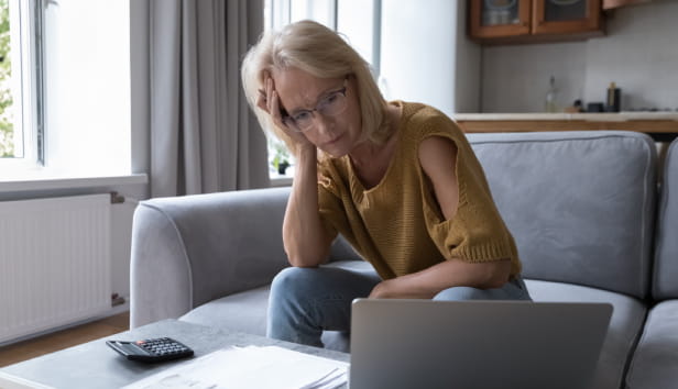 A woman sat on her sofa with her head in her hand. She is staring at a laptop screen