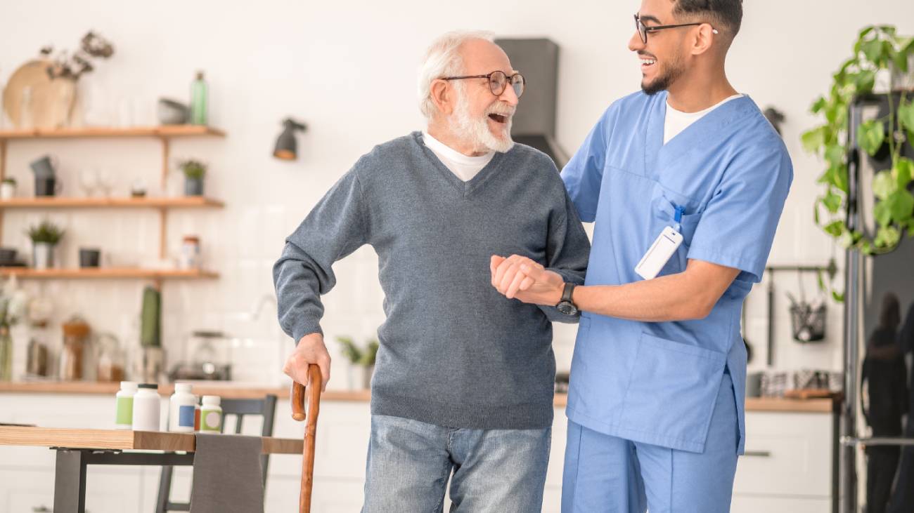 Elderly man walking with a walking stick, aided by a nurse, they are smiling at each other and look very happy in a living room setting 