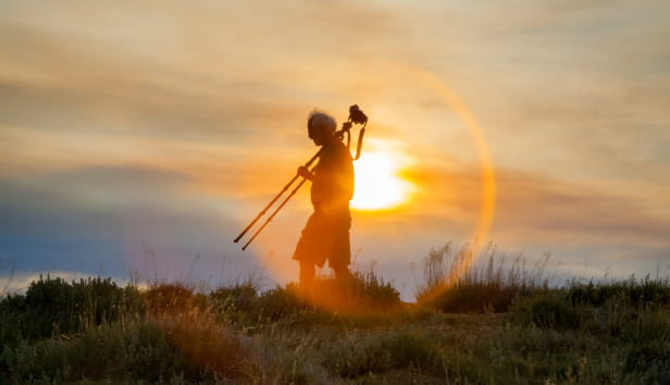 A seasoned photographer with grey hair blowing in the wind, walks at sunset with his tripod resting on his shoulder.  He is in partial silhouette with the sun directly behind him.
