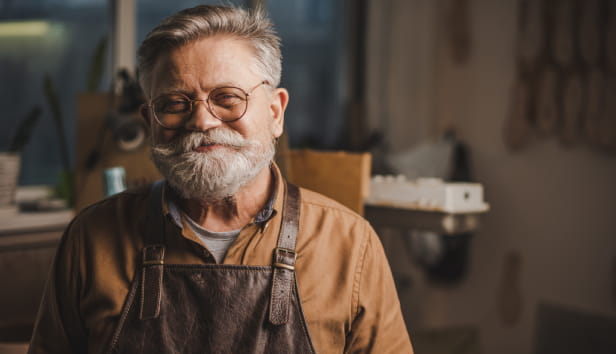 Happy, senior shoemaker in glasses smiling at camera while standing in workshop