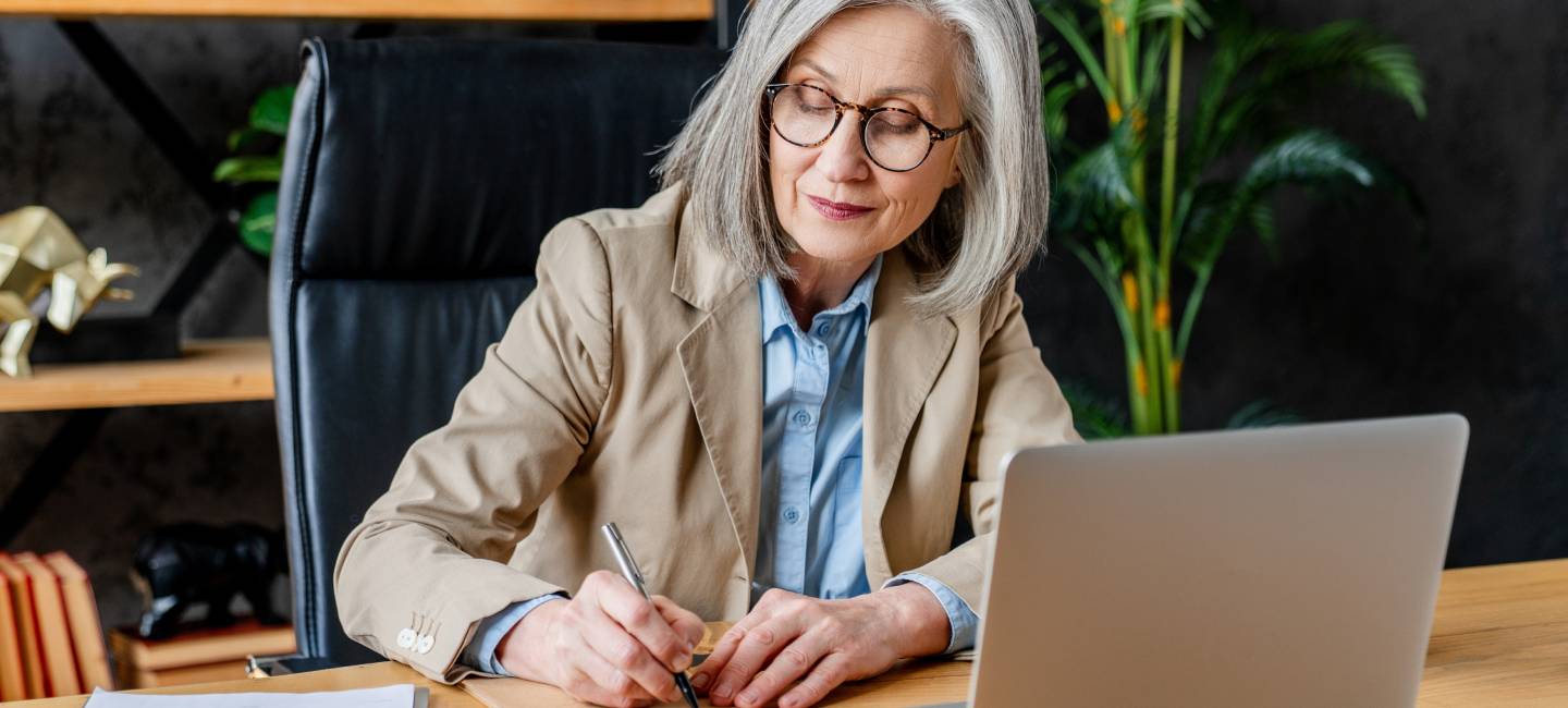 Focused busy mature elderly professional business woman manager executive or entrepreneur using laptop computer in office, working writing notes sitting at desk, modern workplace.