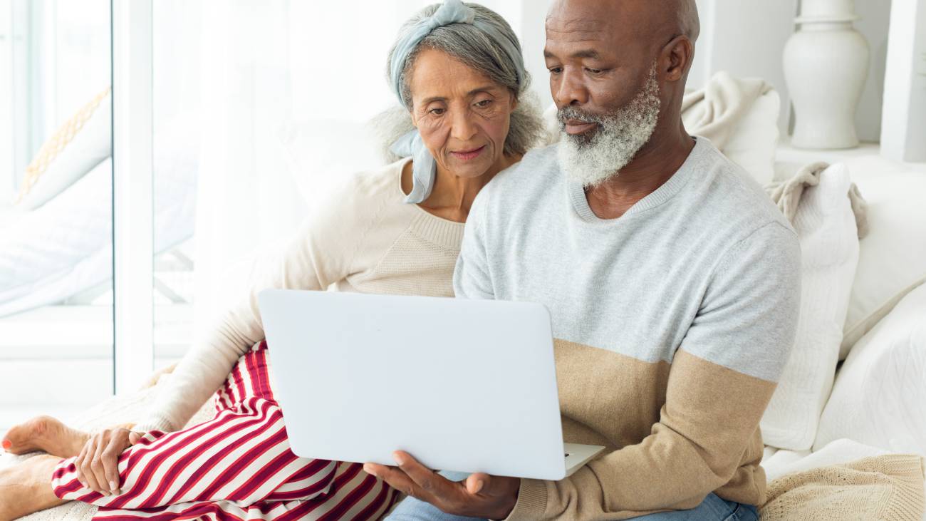 Mature diverse couple sitting on a sofa looking at a silver laptop