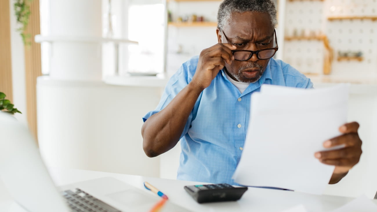 A man sat at a desk looking shocked. There is a laptop, calculator and some papers on the desk