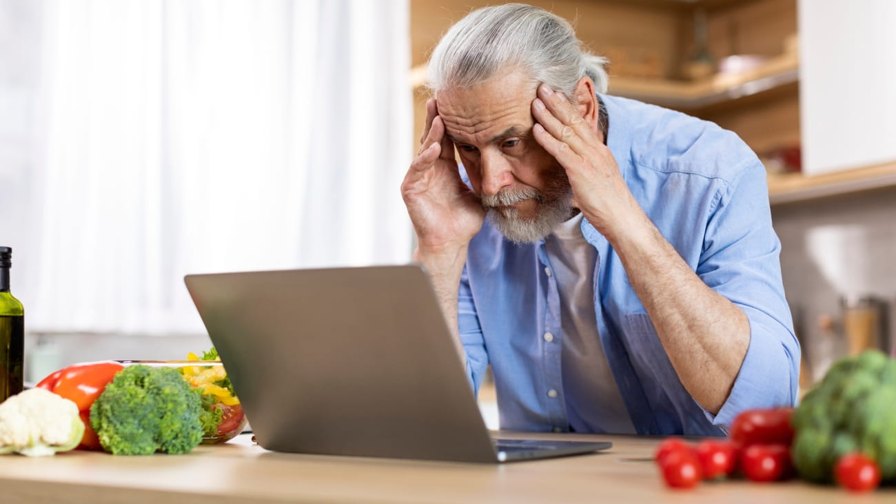 A man sat at his kitchen counter looking at his laptop screen. He has his head in his hands and looks worried