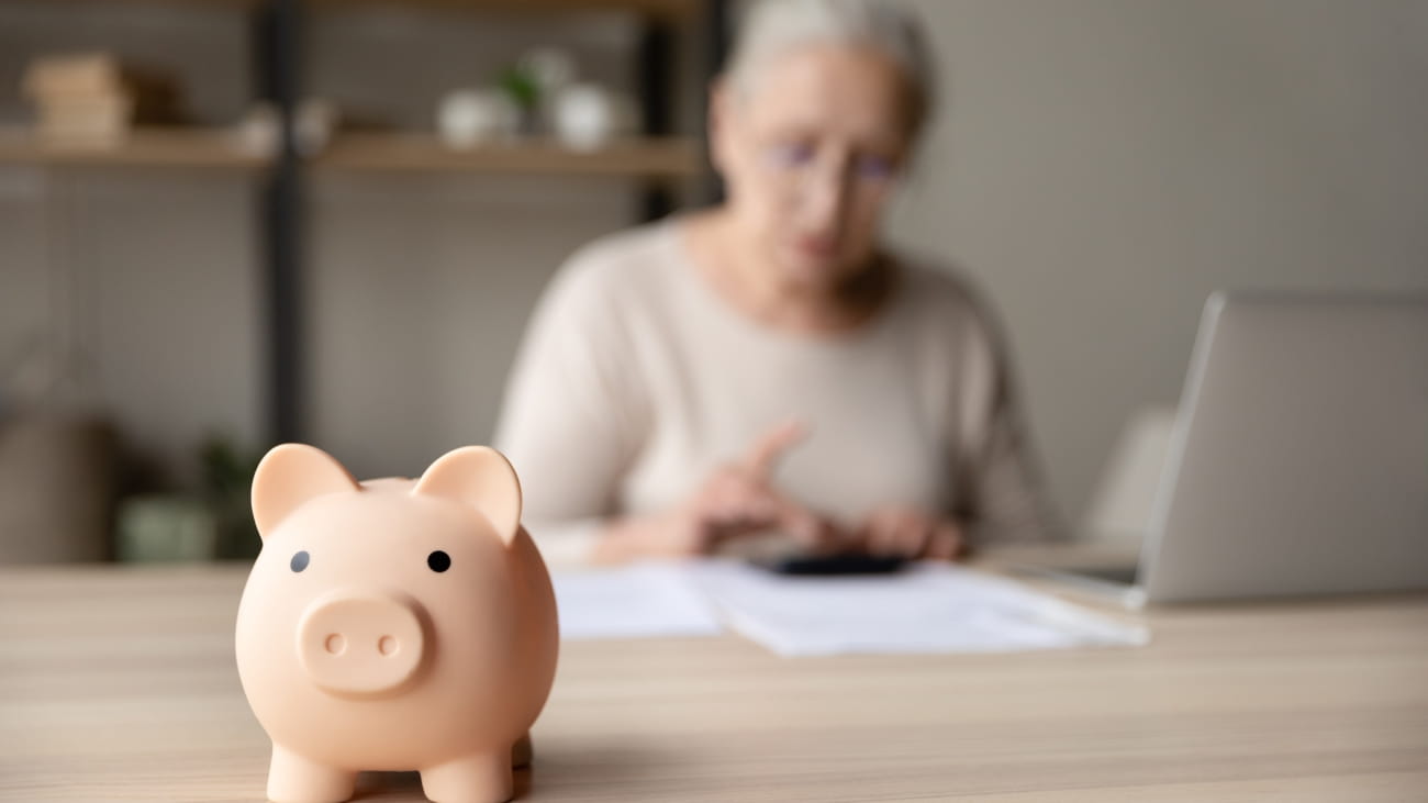 Toy pink piggy bank on work table of senior tenant, homeowner woman. Elderly lady using calculator, counting savings, taxes. Finance management, financial insurance, personal budget concept